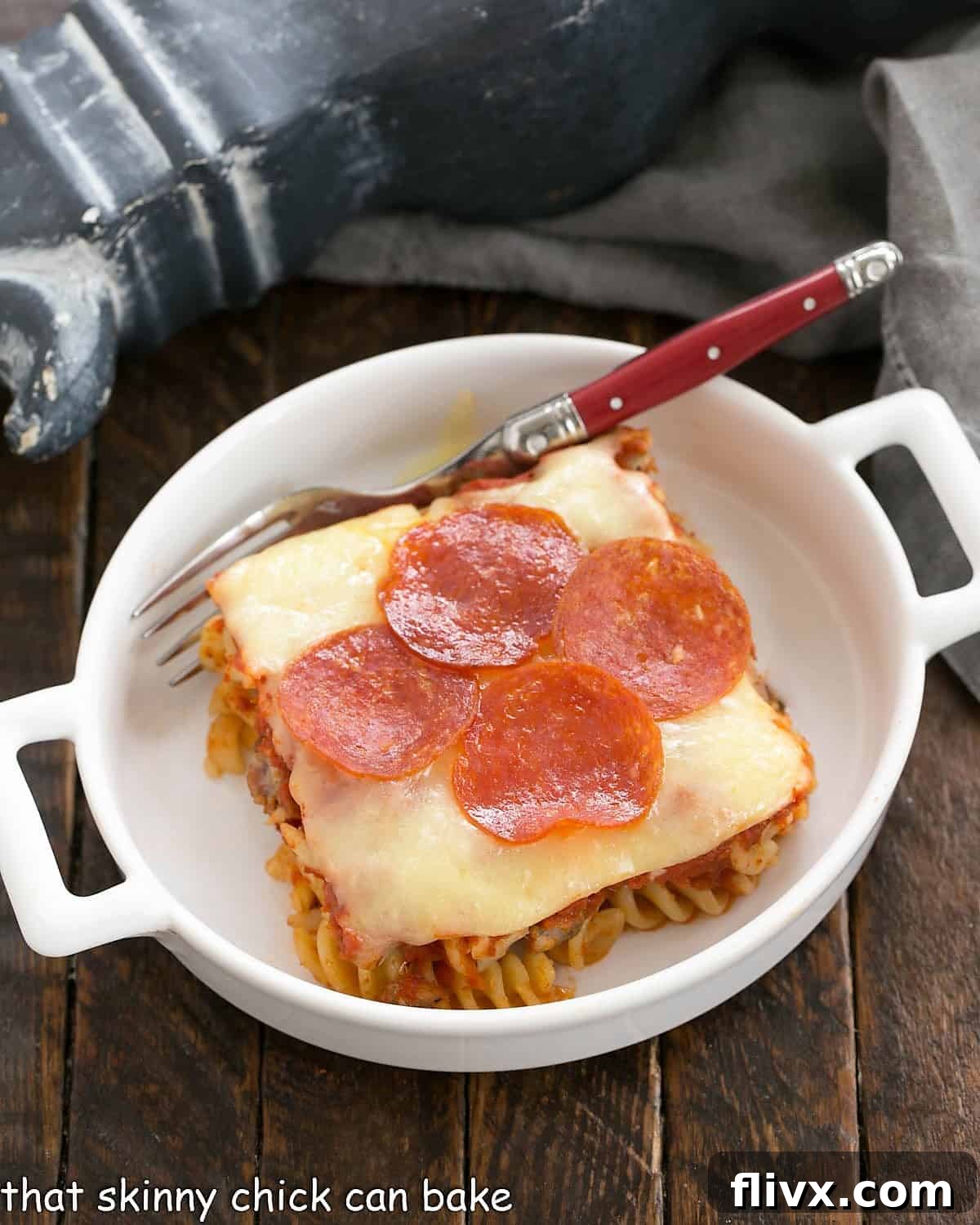 A perfectly sliced portion of Easy Pizza Casserole, served in a white dish with a red-handled fork, showcasing layers of pasta, meat, sauce, and melted cheese.