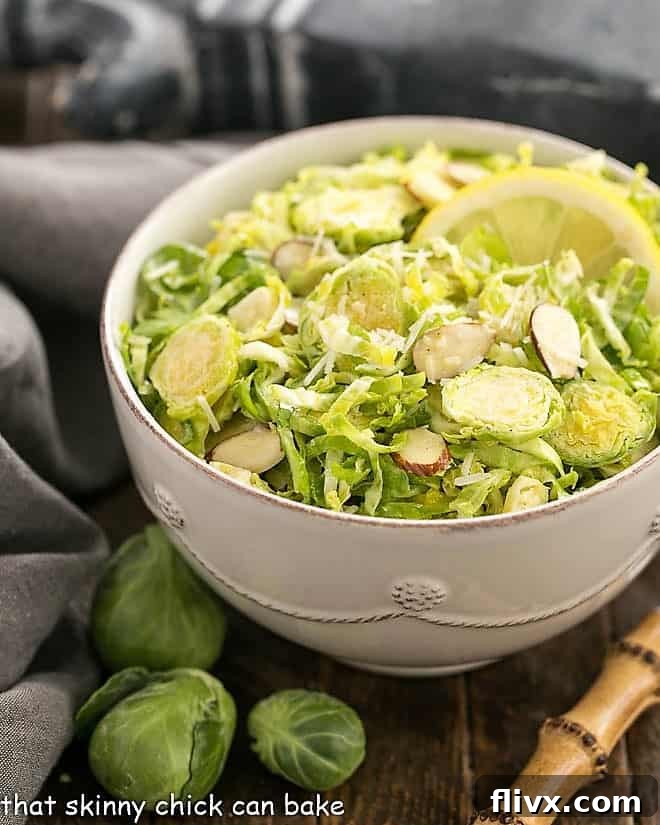 Shaved Brussels Sprouts Salad in a white salad bowl with a lemon slice and a bamboo handled fork, set on a rustic wooden table.