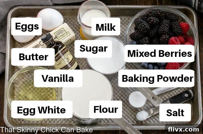 An organized display of ingredients for a berry cake, meticulously arranged on a metal sheet pan. Flour, sugar, eggs, butter, baking powder, milk, and a vibrant assortment of fresh blueberries, raspberries, and blackberries are clearly visible, ready for baking.