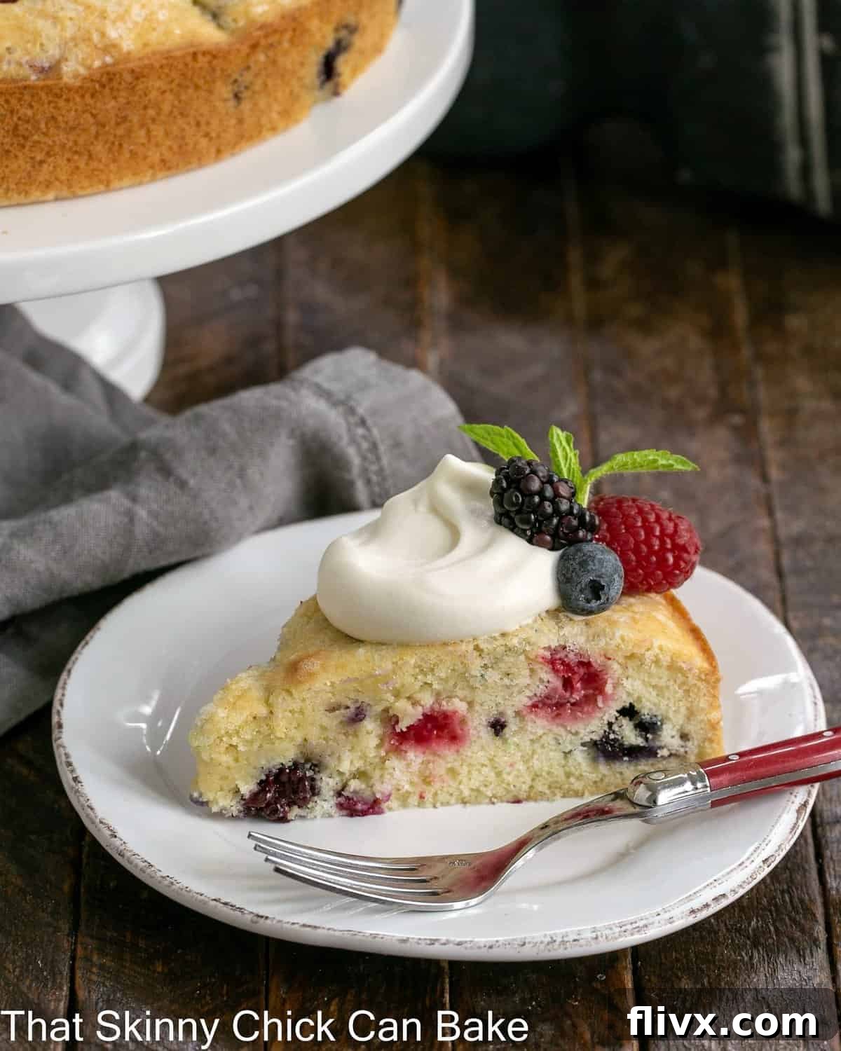 A close-up shot of a perfect slice of berry cake, adorned with fluffy lemon whipped cream, resting on a white plate. In the background, a full cake on a stand hints at the dessert's delightful presentation, emphasizing its light and refreshing quality.