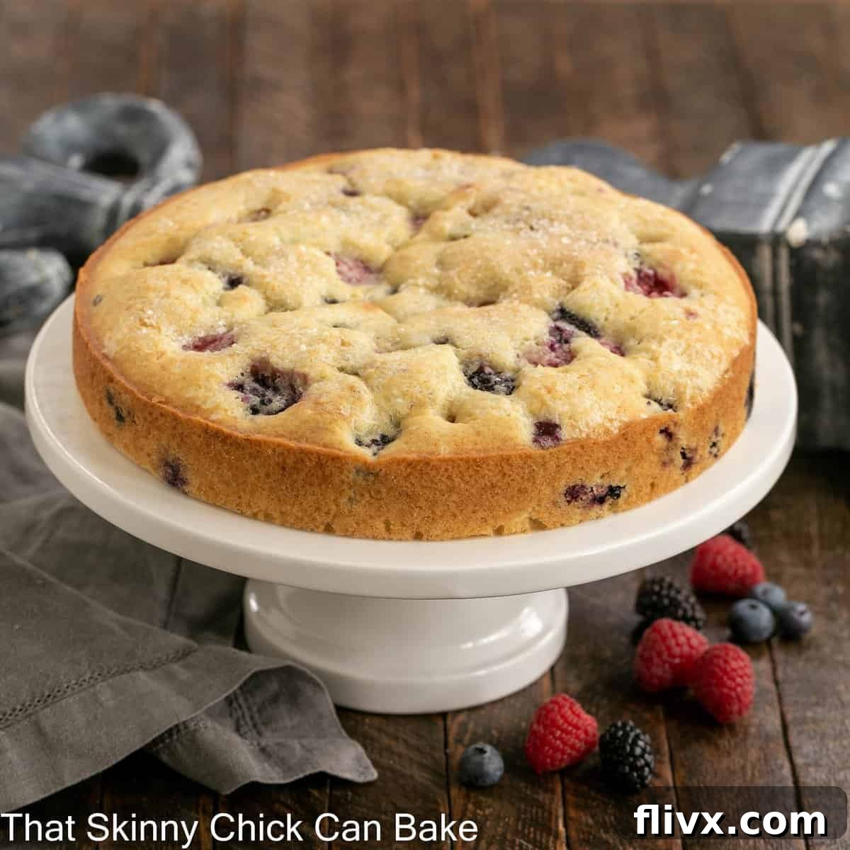A stunning one-layer cake generously studded with mixed berries, proudly displayed on an elegant cake stand. The cake appears golden-brown and perfectly baked, inviting viewers to imagine its fresh, fruity flavor.