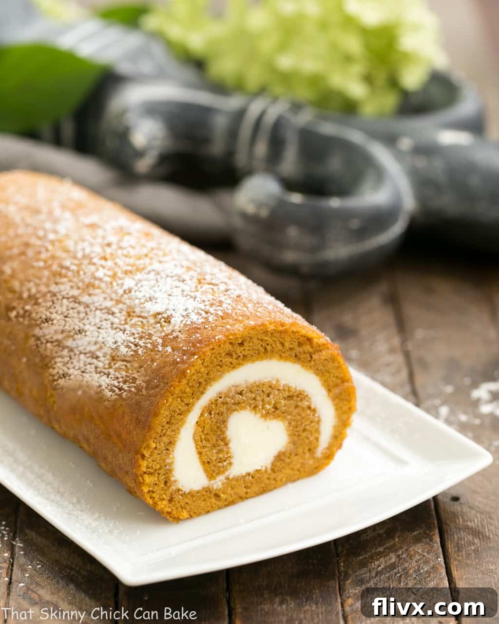 A beautifully presented old-fashioned pumpkin roll with cream cheese filling on a rectangular serving tray, ready for slicing. The rich orange color of the cake contrasts with the snowy white powdered sugar.
