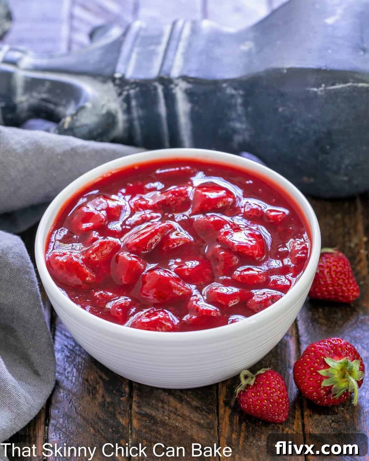 A white bowl full of rich, red strawberry compote with a few fresh, whole strawberries on the side for garnish.