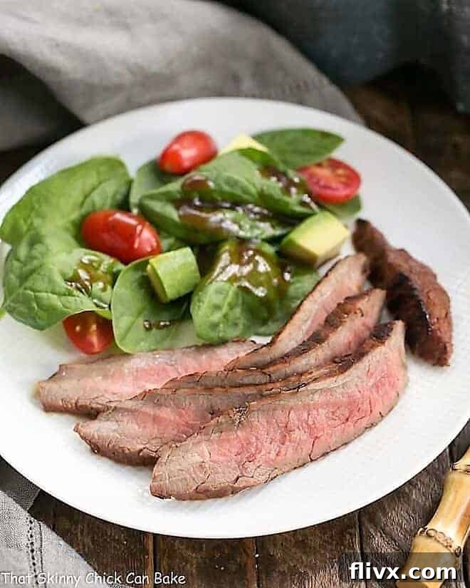 A perfectly cooked, marinated flank steak, with a rich red wine, soy, and balsamic glaze, served on a white dinner plate alongside a fresh spinach salad.
