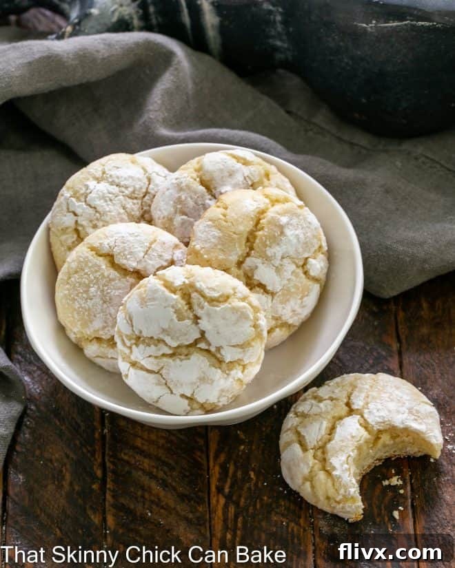 An overhead view of a small white bowl containing five perfectly baked gooey butter cookies, with one cookie placed separately, showing a bite removed to reveal its soft interior.