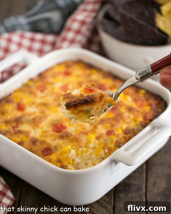 A serving spoon lifting a generous scoop of hot, cheesy Fiesta Corn Dip from a white casserole dish, showcasing its gooey texture.