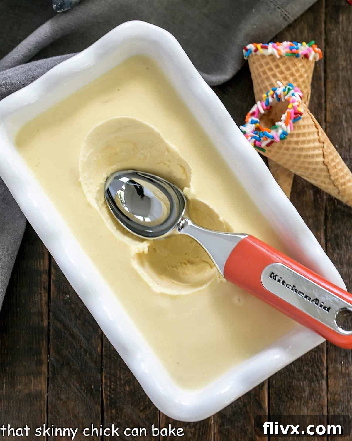Smooth, pale yellow cake batter ice cream resting in a white freezer-safe dish, with a metal ice cream scooper partially submerged, ready to serve.