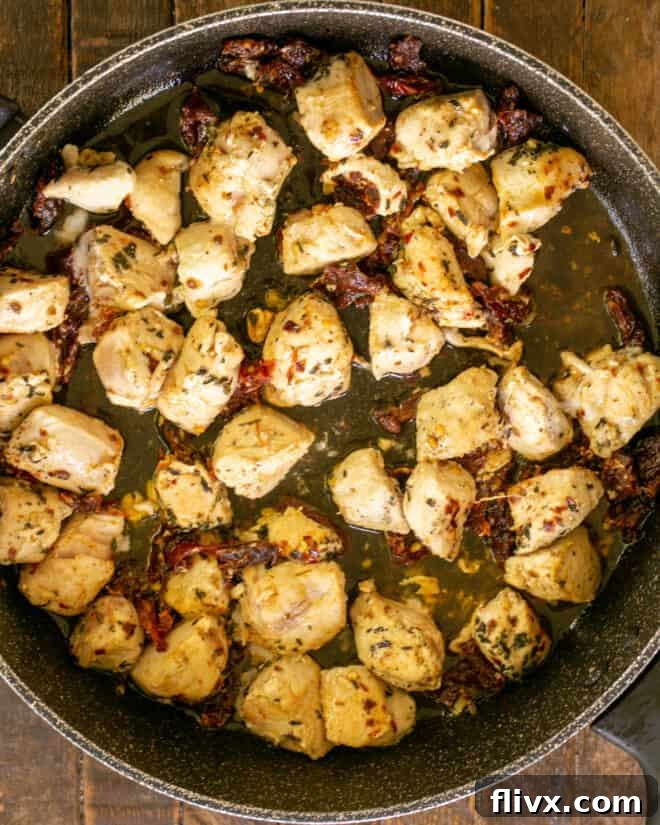 Overhead view of chicken cubes being sautéed in a skillet with minced garlic, dried basil, seasoning salt, and sun-dried tomatoes, creating an aromatic base.