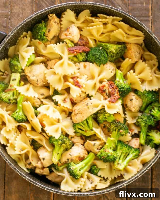 Cooked bow tie pasta being added to the skillet with chicken and broccoli, ready to be tossed and coated in the rich sauce.