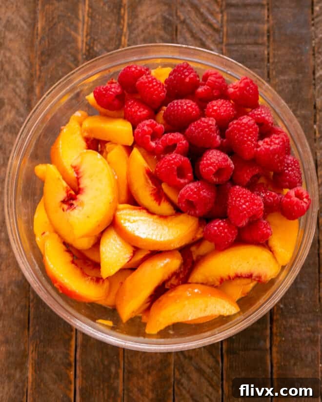 Peeled and sliced peaches combined with fresh raspberries in a mixing bowl, ready for sugar, flour, and cinnamon.