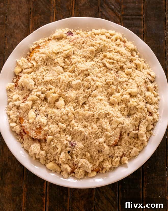 Hands sprinkling the crumbly topping evenly over the peach and raspberry filling in the baking dish, pinching some for larger crumbles.