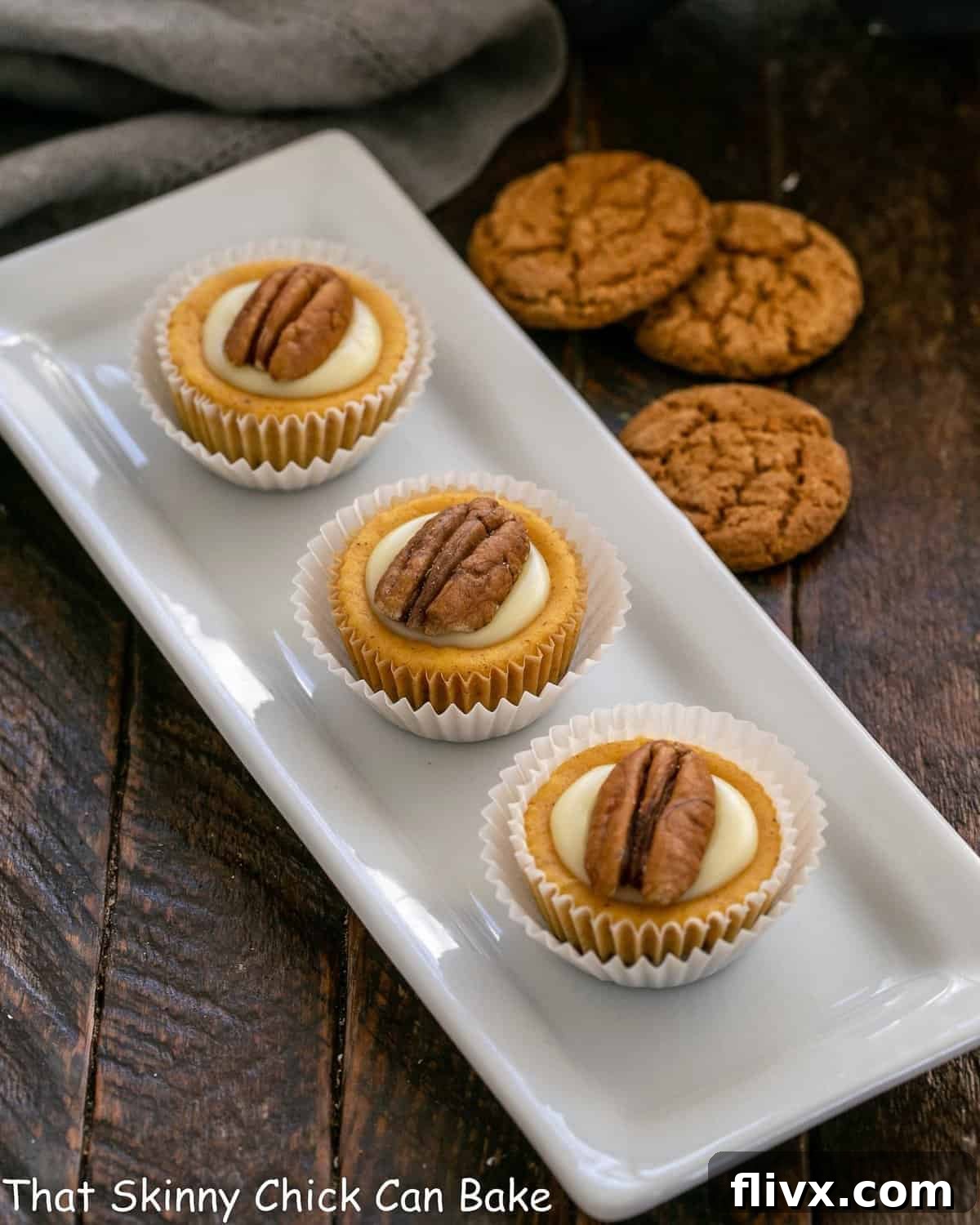 Three perfectly crafted mini pumpkin cheesecakes displayed on an elegant white tray, with gingersnap cookies subtly blurred in the background, hinting at their delicious crust.