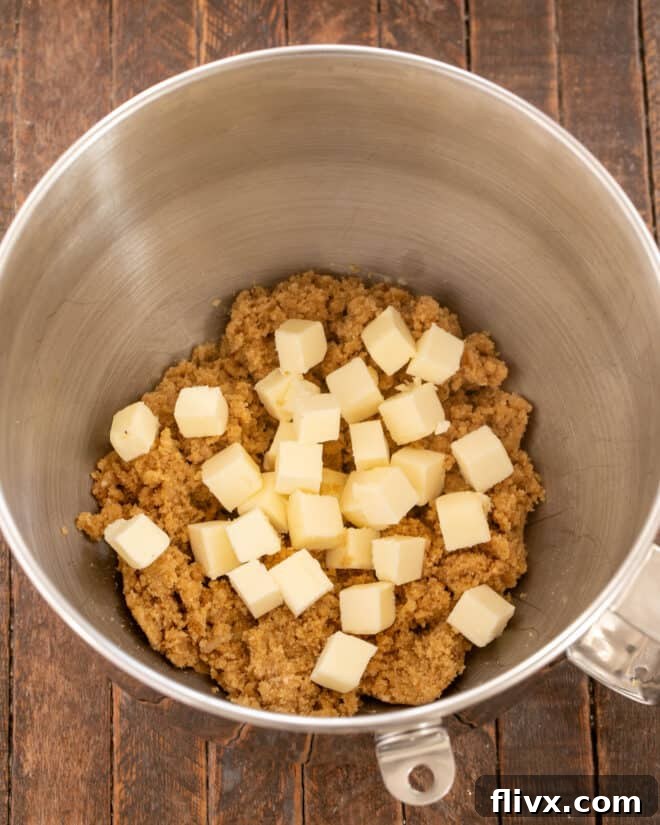 The remaining cold butter pieces being added to the mixer bowl with the creamed butter and sugar mixture.