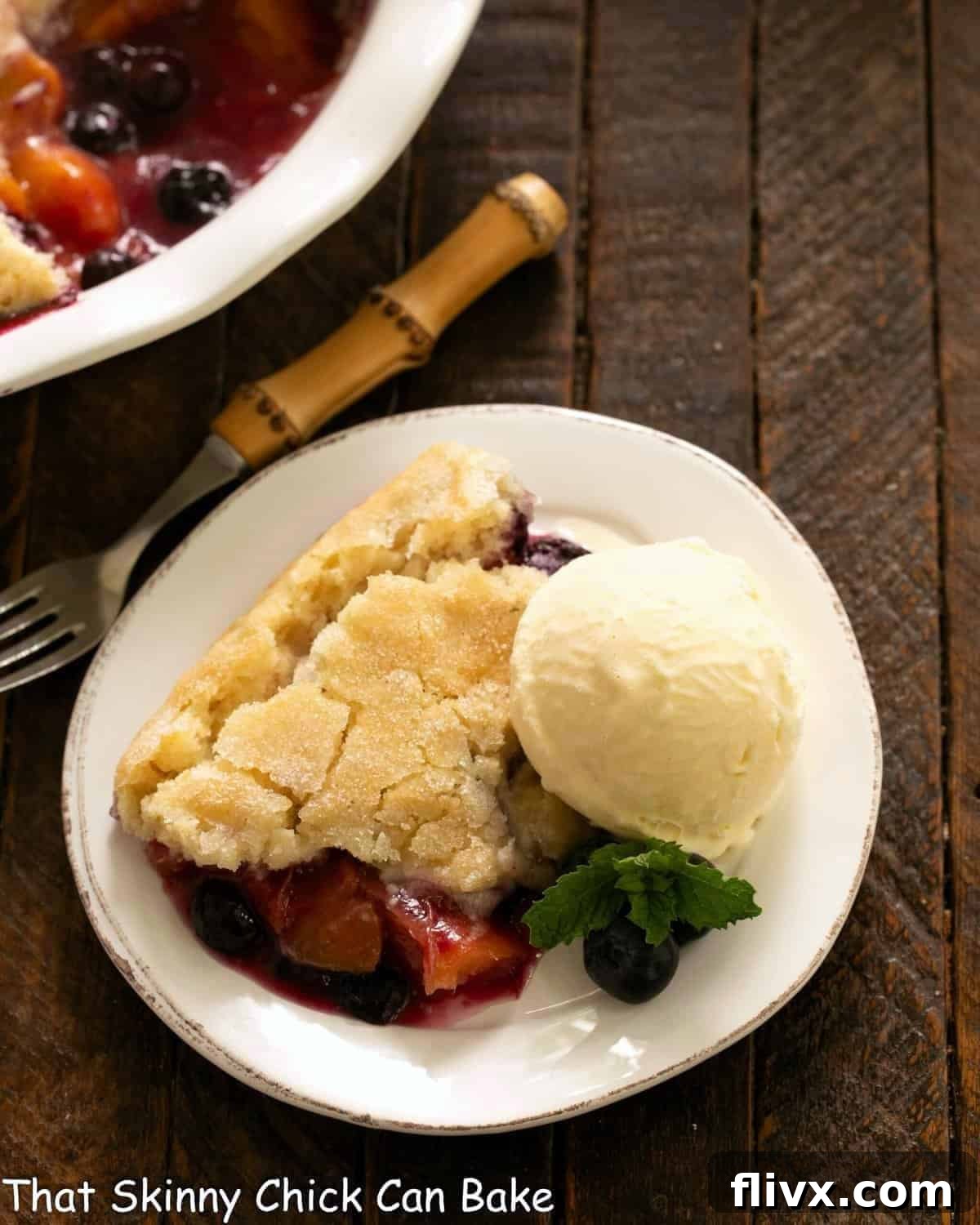 Serving of fruit cobbler with ice cream on a white plate with a fork, emphasizing its warmth and deliciousness.