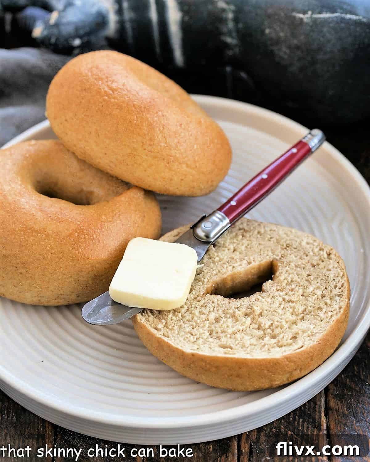 Two golden-brown whole wheat bagels on a white plate, one sliced open with a knife and a pat of butter, showcasing its dense and chewy texture.