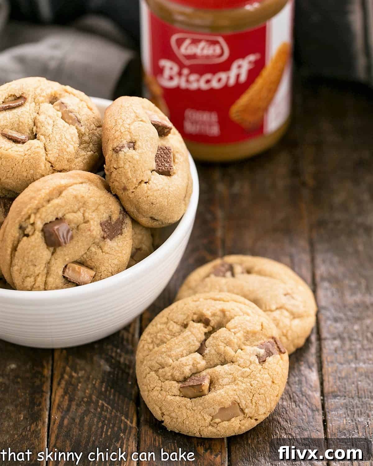 Two perfectly baked Toffee Biscoff cookies positioned next to a white bowl brimming with more cookies, with a jar of cookie butter in the background.