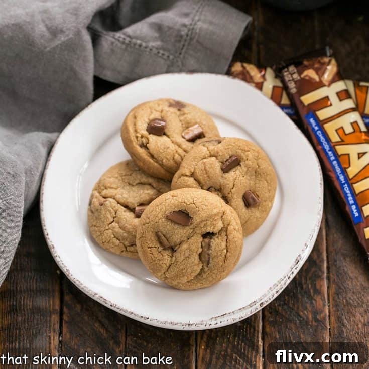 Overhead view of several perfectly baked Biscoff Toffee Cookies on a white plate, alongside two Heath bars, emphasizing the key ingredients.