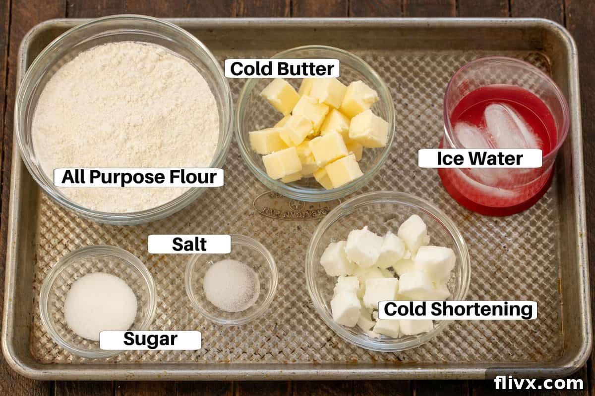 All the necessary ingredients for making a pie crust, including flour, cold butter, shortening, salt, sugar, and ice water, neatly arranged on a baking sheet.