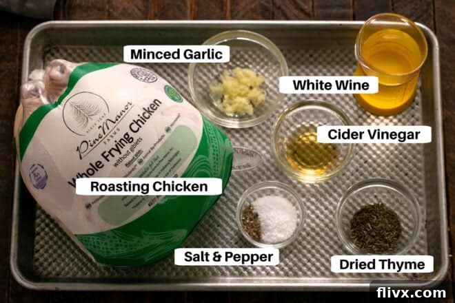 All the essential ingredients for a Whole Roasted Chicken laid out on a metal sheet pan: a whole raw chicken, fresh garlic cloves, a small bowl of dried thyme, cider vinegar, and a bottle of dry white wine. Each ingredient is perfectly positioned for easy identification.