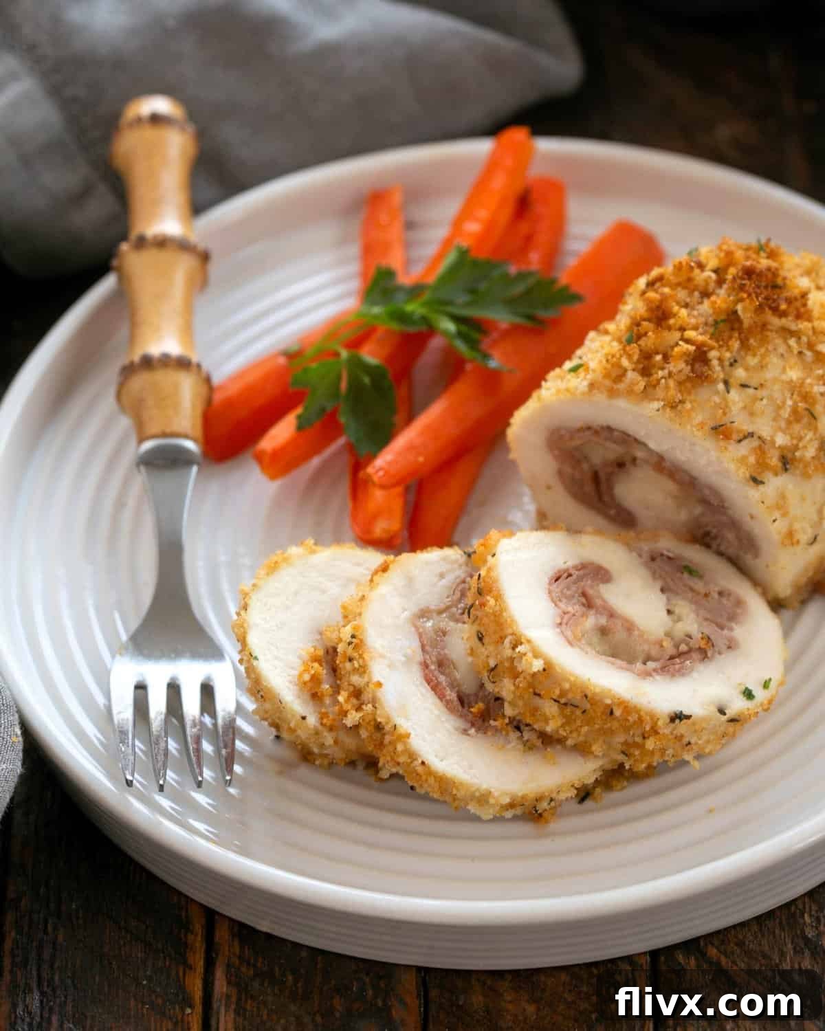 A perfectly baked Chicken Cordon Bleu, partially sliced to reveal the melted cheese and ham, served on a white plate with a side of vibrant carrots and a fork.