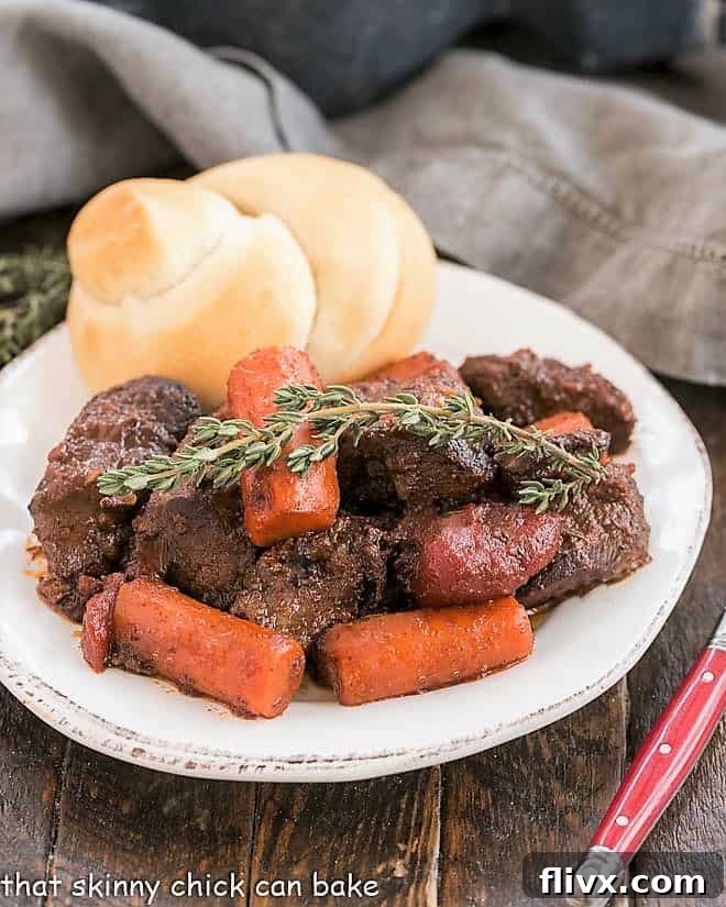 Beef stew with carrots and a sprig of thyme with a dinner roll on a white plate.