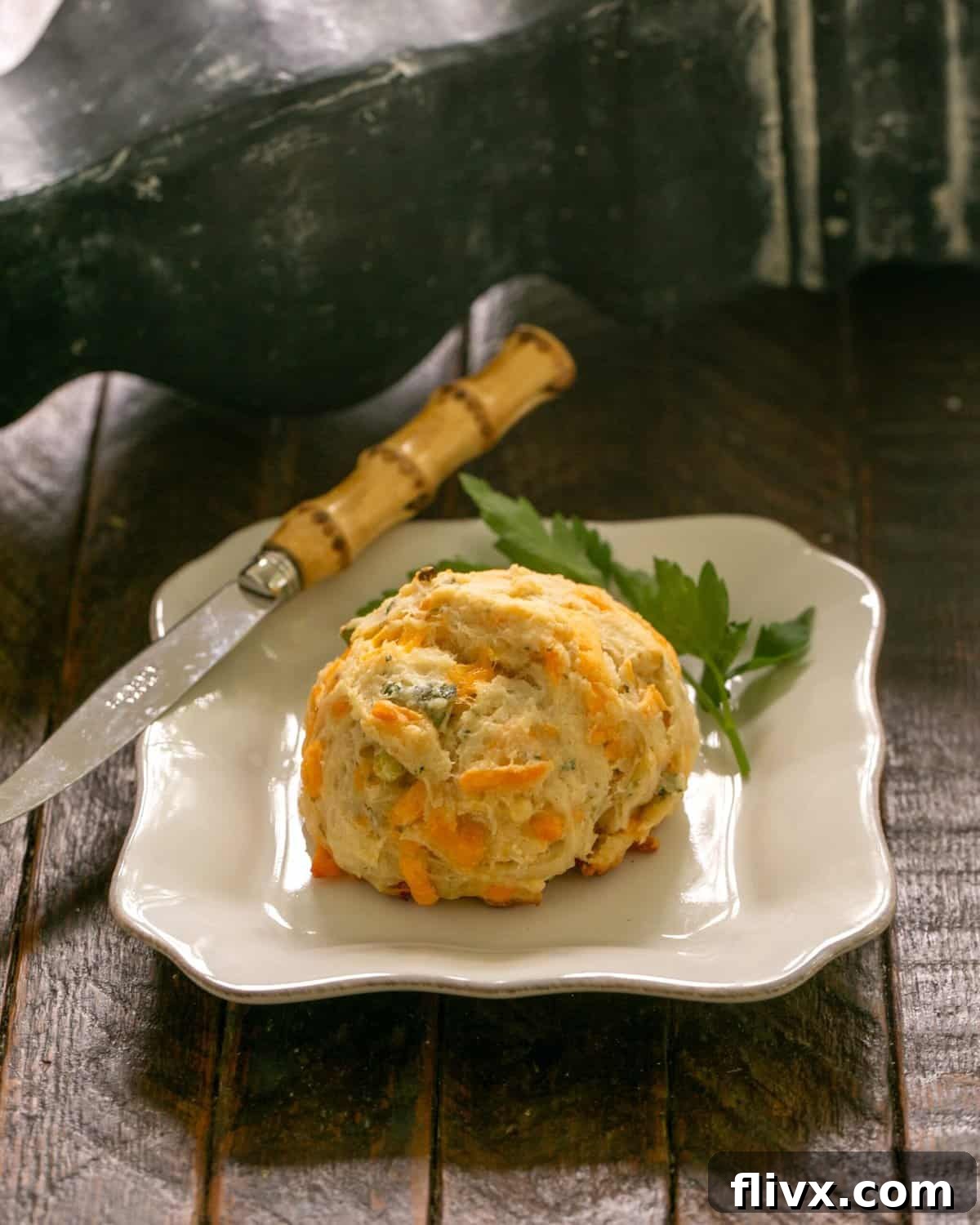 A golden-brown cheddar biscuit on a white plate, garnished with a fresh sprig of parsley, with a butter knife beside it.