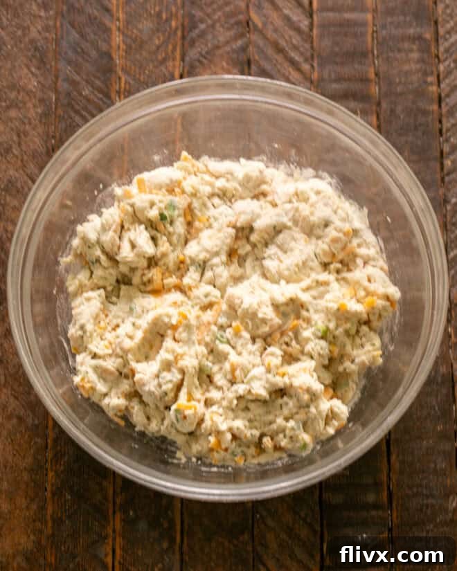 Shredded cheddar cheese, sliced green onions, and chopped fresh parsley are being added to the bowl of whisked dry ingredients, followed by buttermilk.