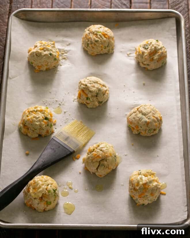 Scoops of cheddar biscuit dough are neatly arranged in quarter-cup mounds on a baking sheet lined with parchment paper.