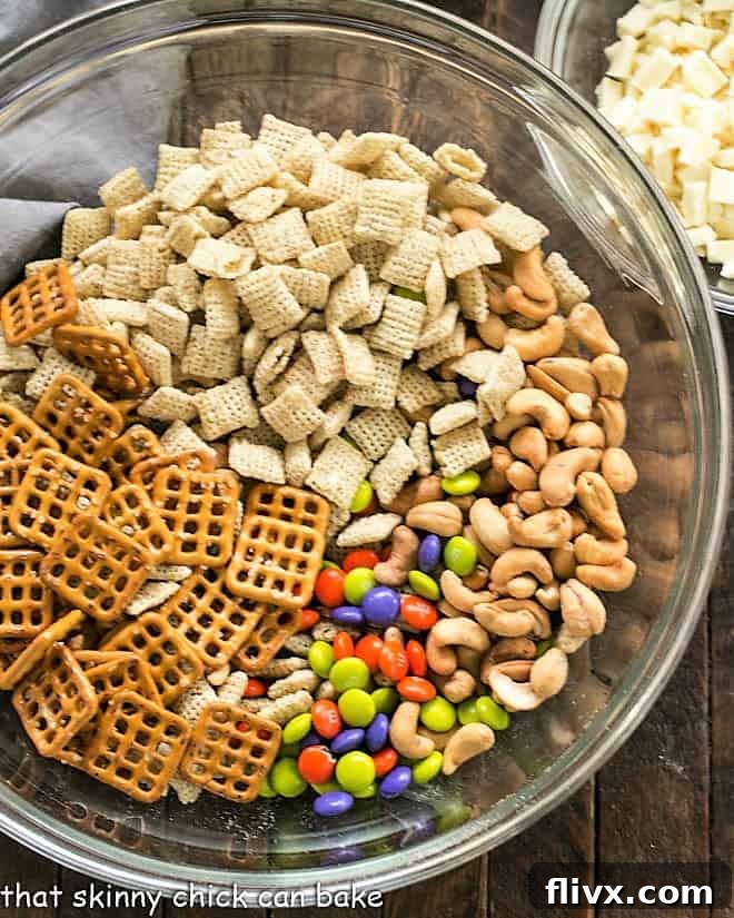 Bowl of various ingredients for White Chocolate Chex Mix, including cereal, nuts, pretzels, and M&M's, ready to be coated.