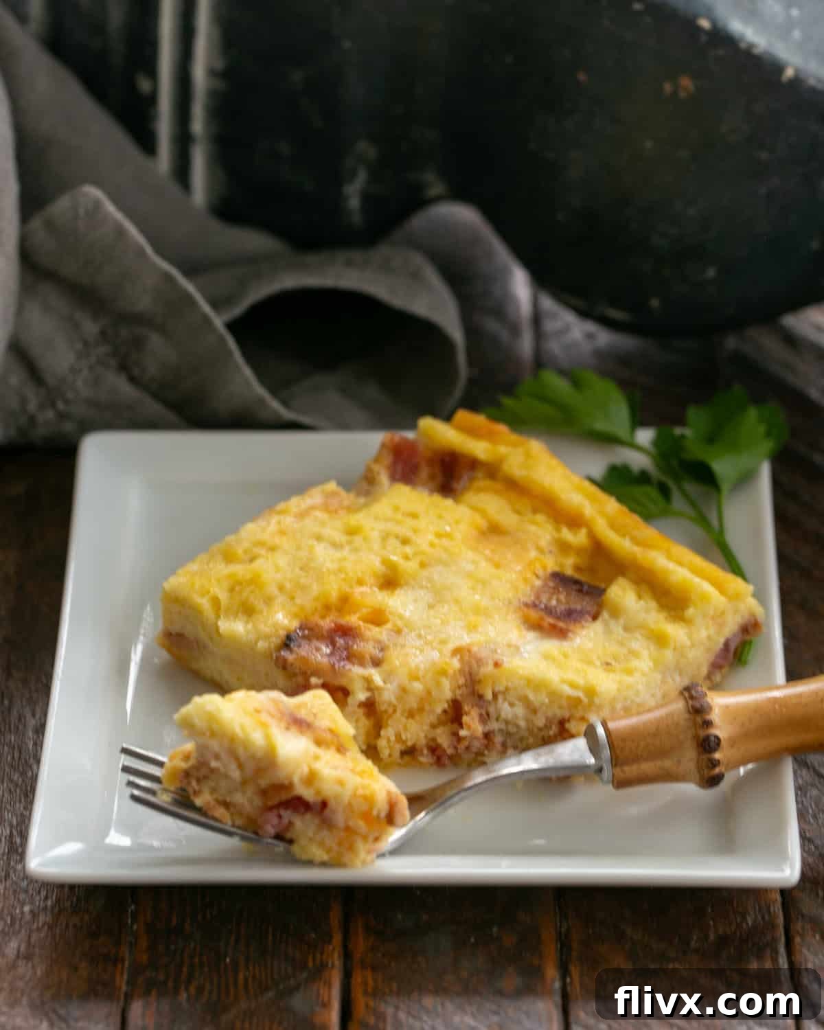 A serving of bacon and egg casserole on a white plate with a fork, garnished with fresh herbs.