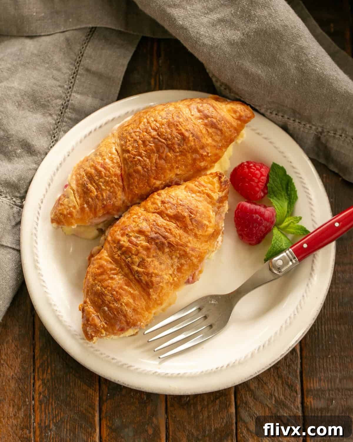 Two golden-brown Croissant French Toast slices, filled with cream cheese and raspberries, garnished with fresh raspberries and powdered sugar on a white plate.