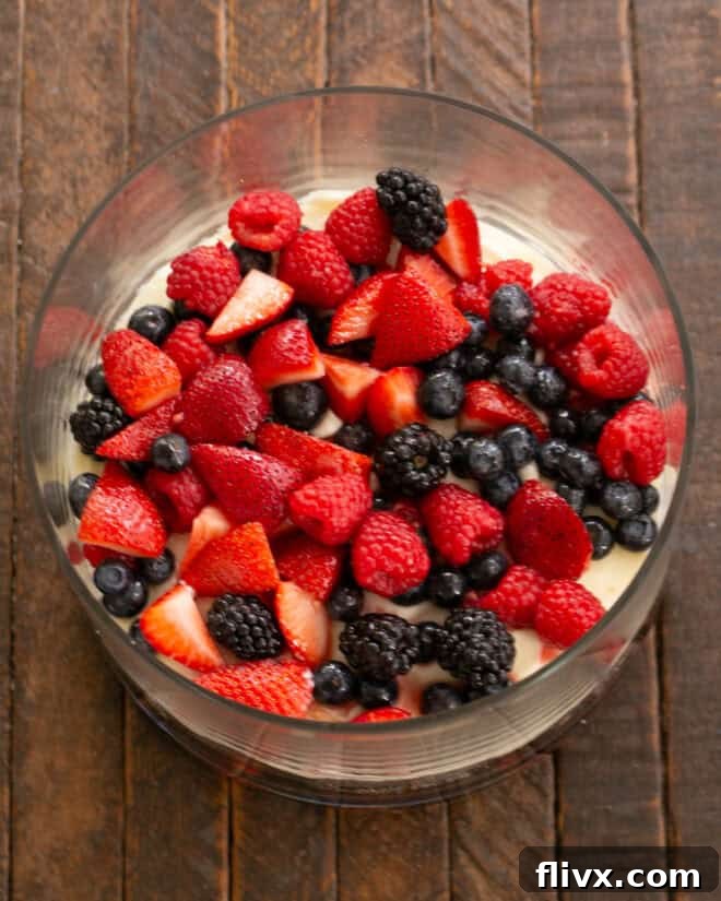 Mixing vibrant fresh berries with sugar and a hint of Amaretto in a bowl. Following this, demonstrating the alternating layers of fluffy cubed angel food cake, the sweetened berry mixture, and the creamy cheesecake filling within a clear trifle bowl.