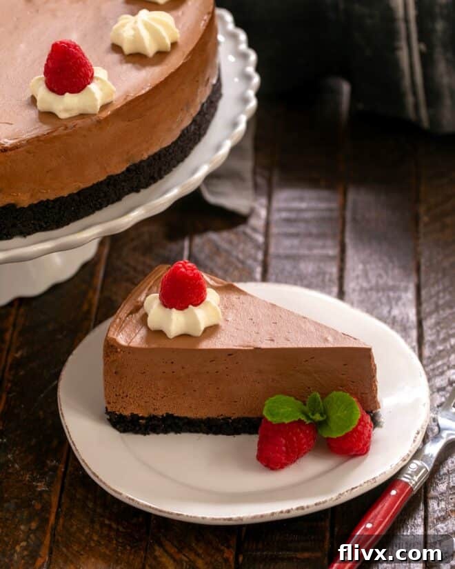 A perfectly sliced piece of chocolate mousse cake on a white dessert plate in front of a full cake on a stand, garnished with berries.