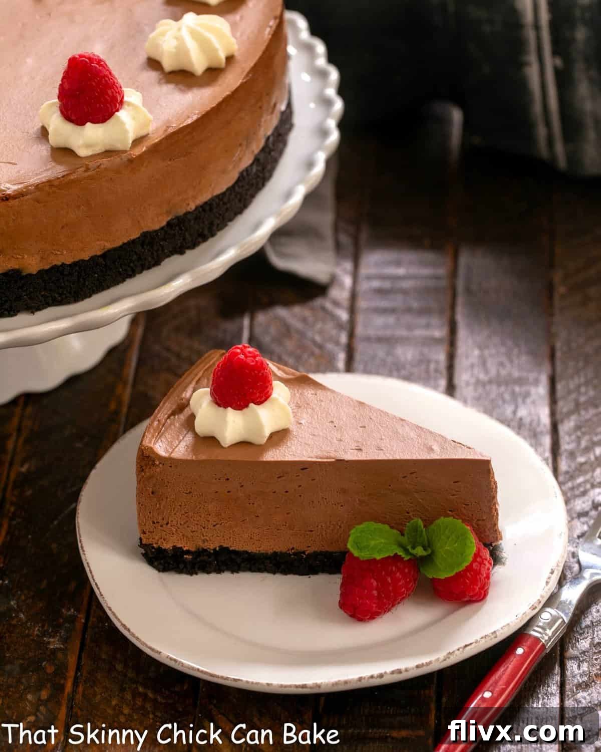 A slice of mousse cake on a white dessert plate in front of a cake stand, garnished with berries and chocolate leaves.