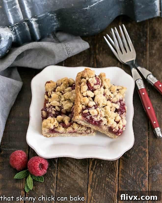 Two vibrant Raspberry Crumble bars resting on a white dessert plate, accompanied by two elegant red-handled forks, ready to be enjoyed.