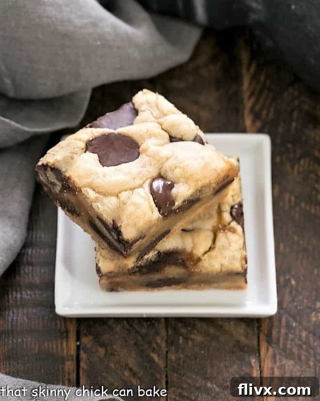 Two golden-brown chocolate chip cookie bars with visible caramel peeking through, stacked on a small square white plate.