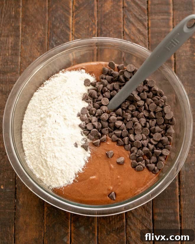 The dry flour mixture and additional chocolate chips or chunks are being added to the rich chocolate cookie batter in a mixing bowl, preparing for the final blend before chilling.