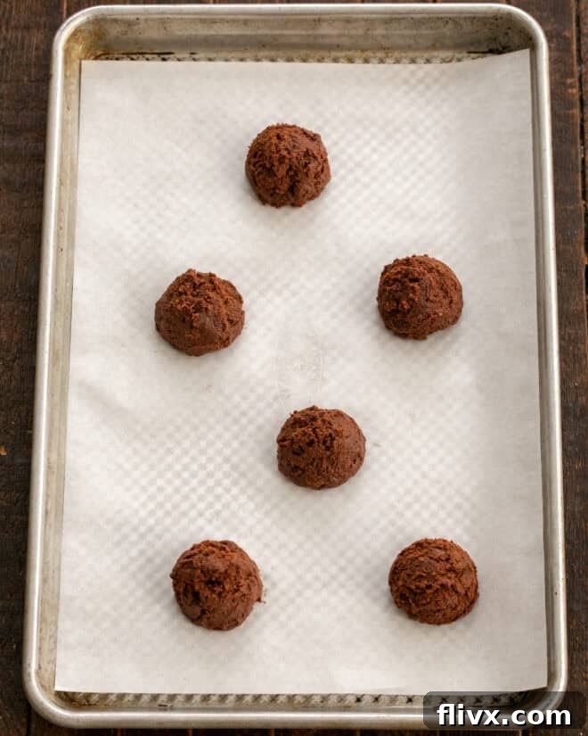 Scoops of chilled chocolate cookie dough are neatly arranged on a prepared baking sheet, spaced adequately apart, ready to be baked into delicious double chocolate cookies.