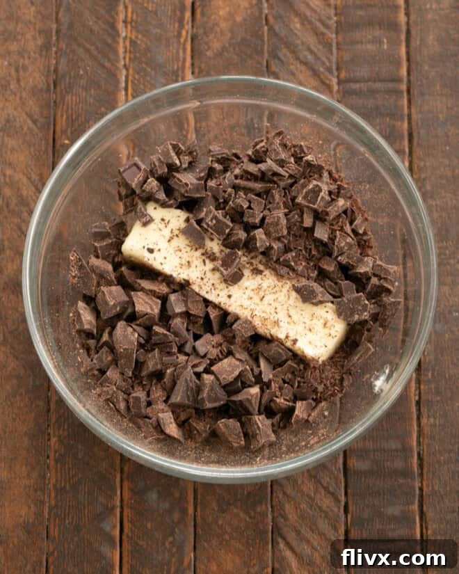 Chopped butter and various types of chocolate chunks are carefully placed into a heat-safe mixing bowl, preparing for the melting process to create the base of the rich double chocolate cookie batter.