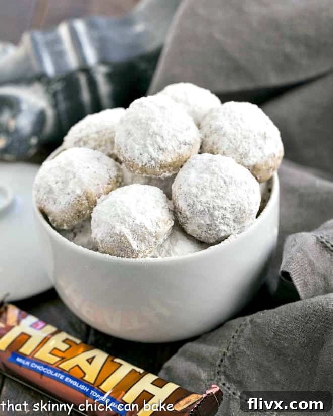 Close-up of Mexican Wedding Cookies with Toffee bits, glistening with powdered sugar, nested in a white serving bowl, ready to be enjoyed.