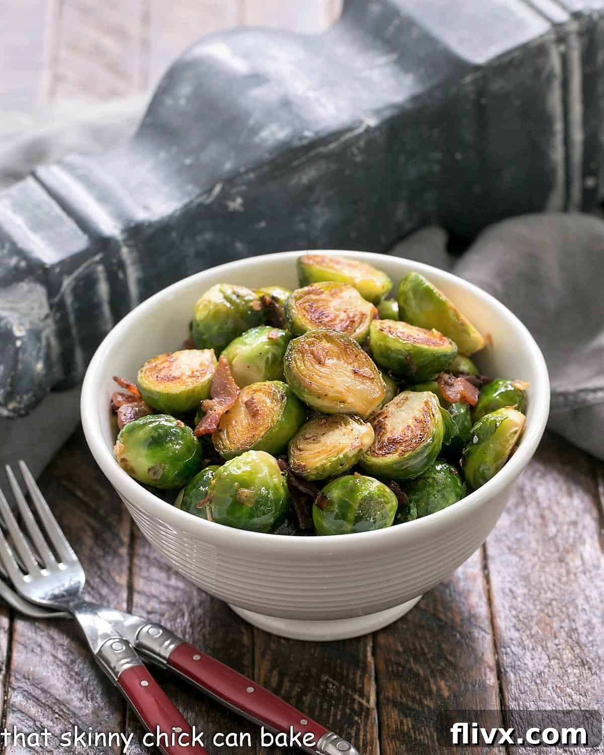A white bowl of skillet brussels sprouts with bacon and two red handled forks.