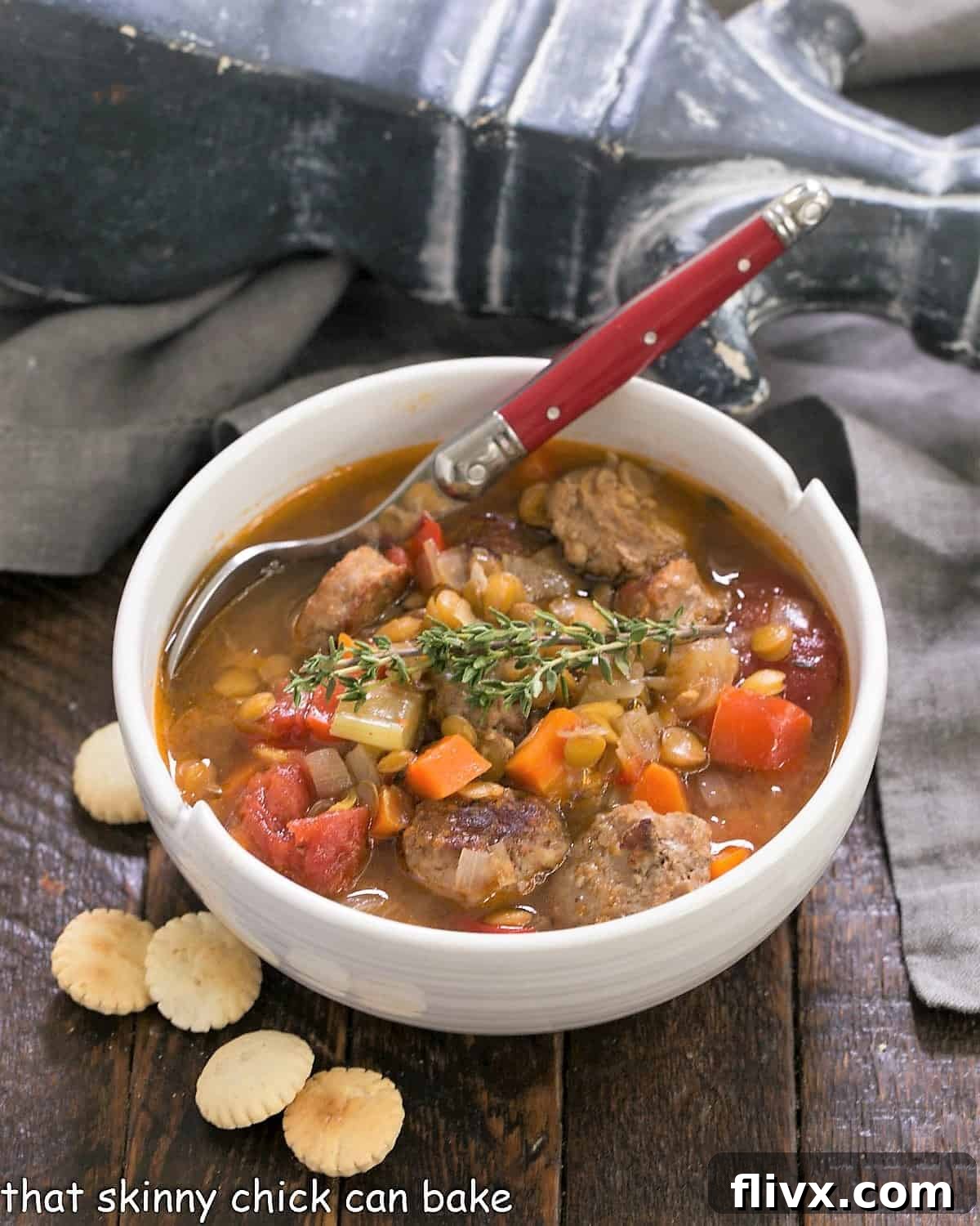 White bowl filled with delicious Sausage Lentil Soup, garnished with fresh herbs and served with crackers and a red handled spoon.