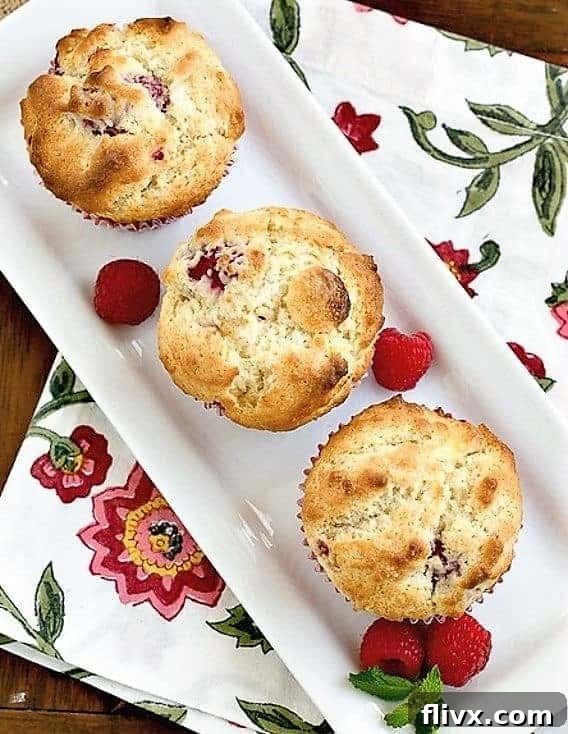 Overhead view of three perfectly baked Raspberry White Chocolate Muffins on a rectangular tray, garnished with fresh raspberries and a sprig of mint.