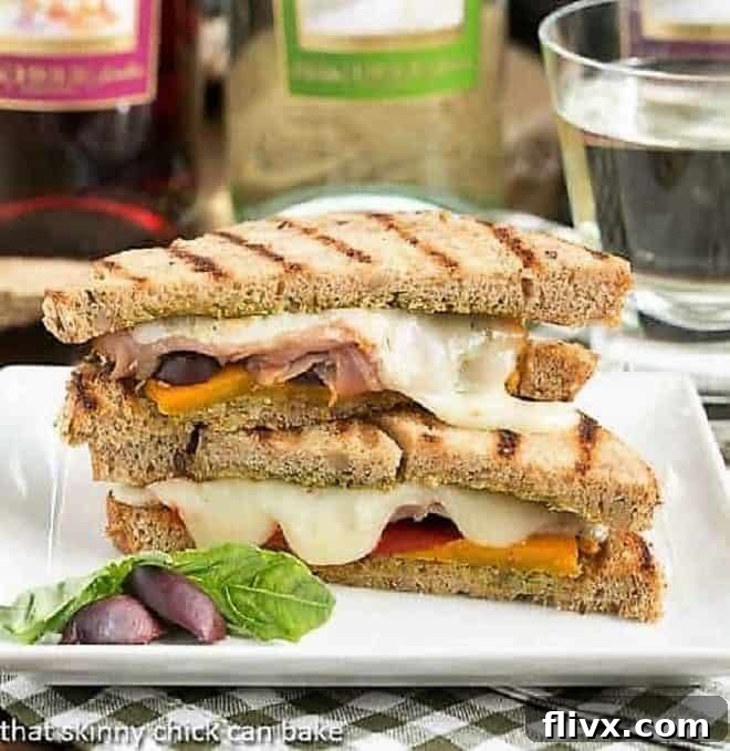 A perfectly sliced Mediterranean Grilled Cheese sandwich, revealing layers of melted cheese, vibrant roasted red peppers, salty prosciutto, and dark Kalamata olives, plated on a clean white dish with a basil garnish.