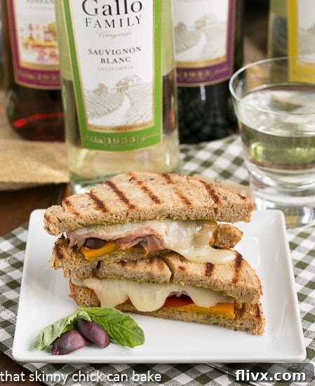 A Mediterranean Toasted Cheese Sandwich, cut in half to show the melted cheese and colorful fillings, elegantly presented on a square white plate with a fresh basil leaf and an olive garnish. 