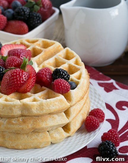 A close-up of Homemade Buttermilk Waffles stacked on a white ceramic plate with a trio of fresh berries (strawberries, raspberries, blueberries).