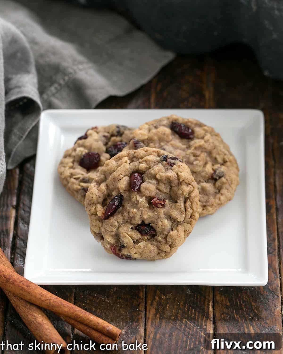 Three golden-brown Oatmeal Craisin Cookies artfully arranged on a square white plate, accompanied by two fragrant cinnamon sticks, highlighting their irresistible texture and vibrant cranberry flecks.