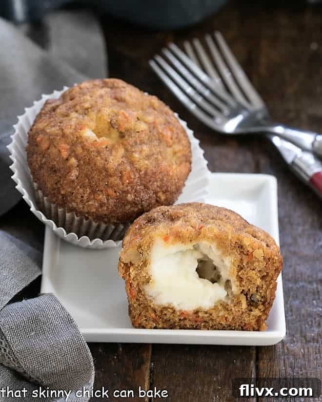 A beautifully baked carrot cake muffin, cut in half to reveal its smooth, white, creamy cheese center, elegantly presented on a square white plate.