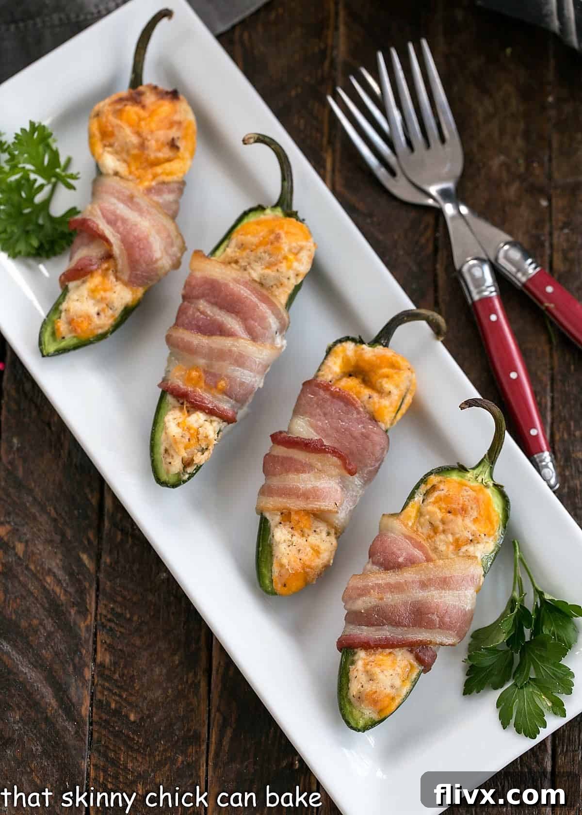 An overhead view of four perfectly baked bacon-wrapped jalapeno poppers arranged on a clean white tray, accompanied by two red-handled forks, inviting you to dig in.