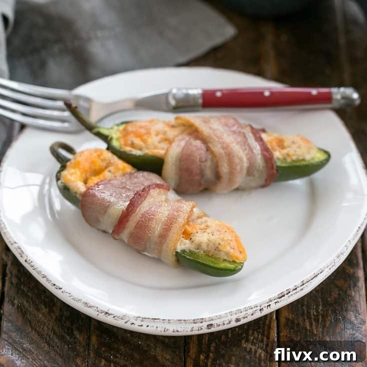 Two bacon wrapped jalapeno poppers on a white ceramic plate with a red handled fork, highlighting their crispy bacon and creamy filling.